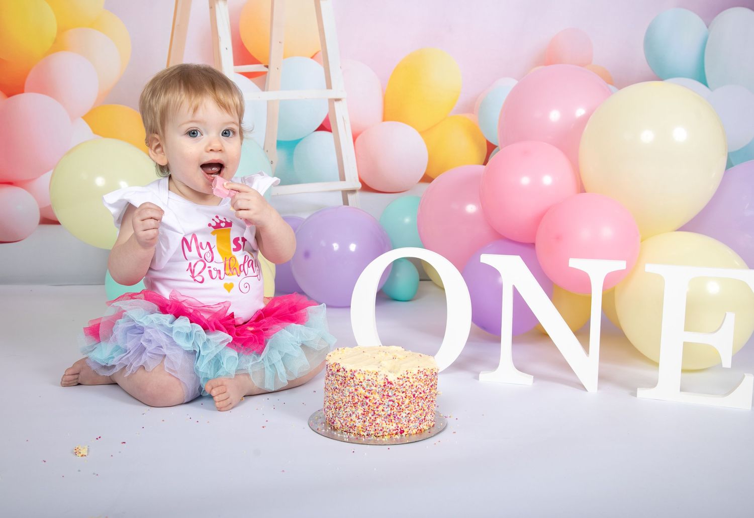 Festive first birthday celebration setup with pastel balloons, ONE letters, and a decorative sprinkle cake.