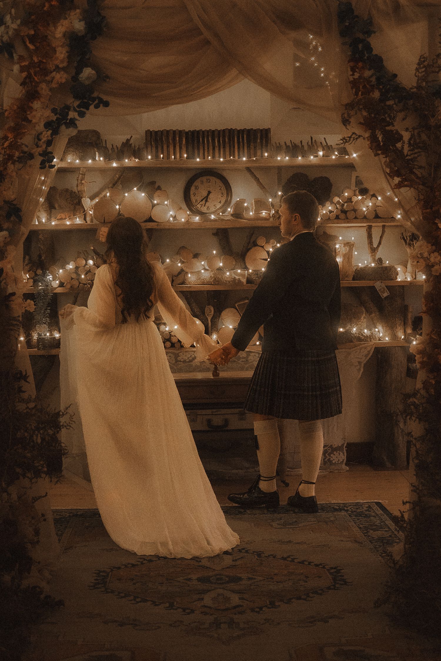 A romantic wedding scene in warm sepia tones showing a couple holding hands in front of a candlelit mantle decorated with greenery.