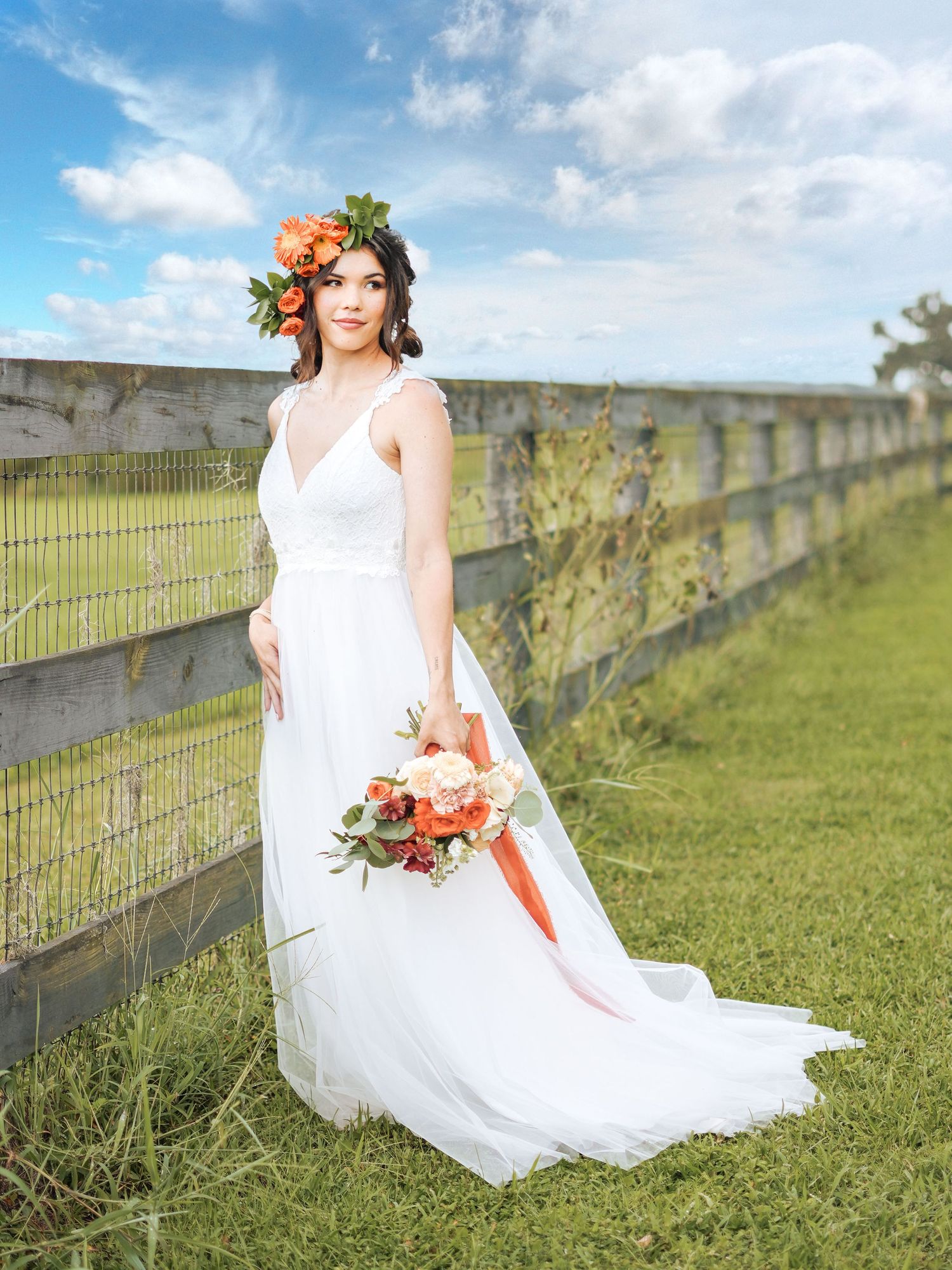 Bride in white gown with coral flower crown poses against blue sky and wooden fence.
