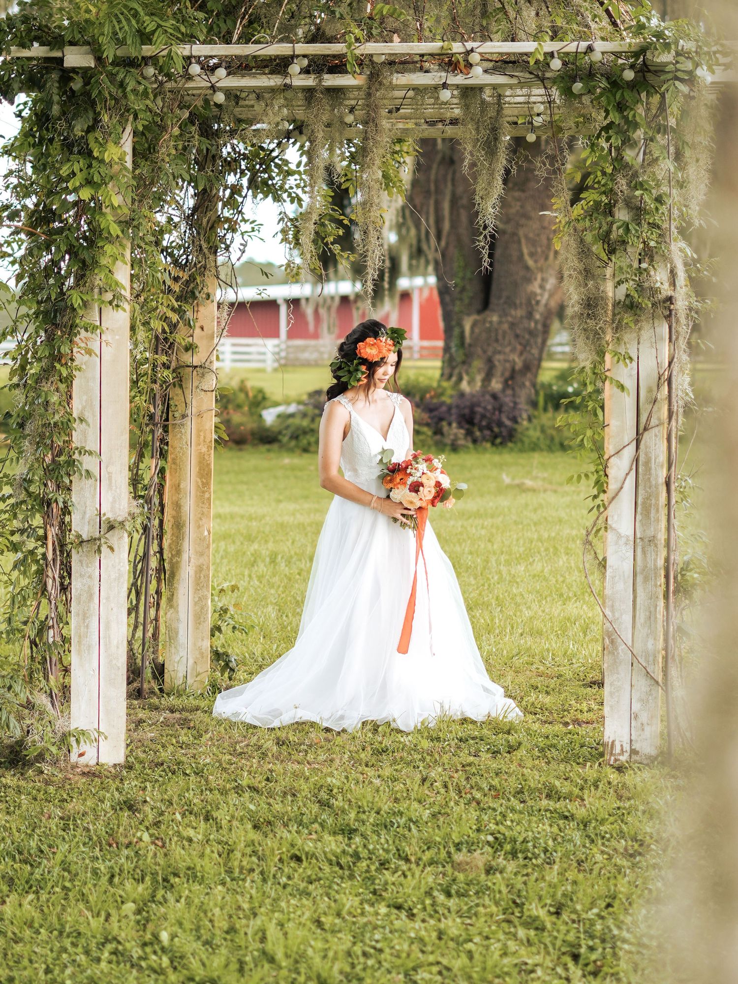 A bridal photo under a vine-covered pergola featuring a white wedding dress and coral bouquet in a garden setting.
