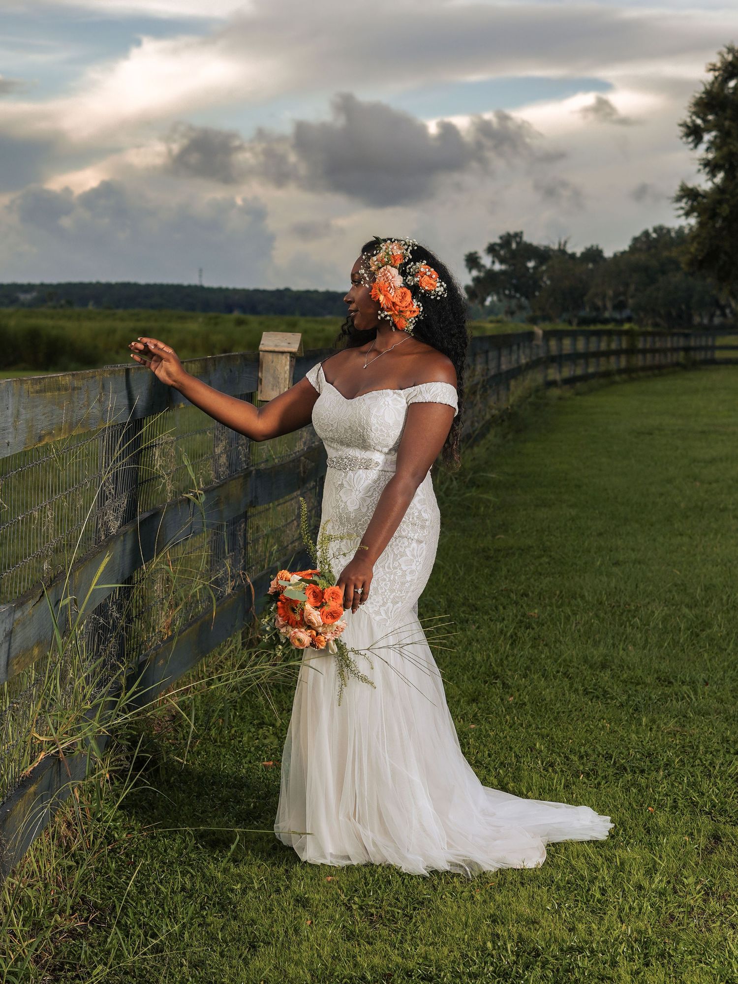 Dreamy bridal portrait with coral florals and white gown against a countryside fence at golden hour.