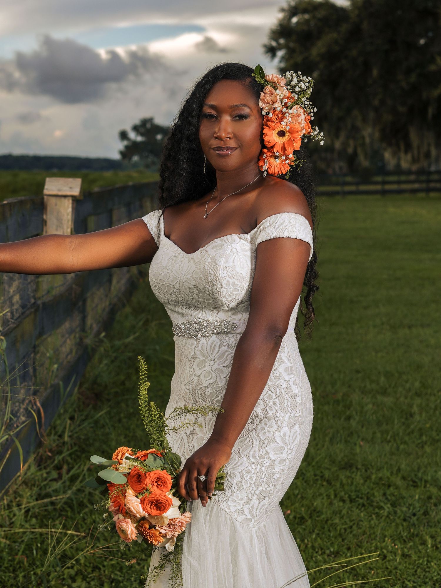Romantic bridal portrait featuring a fitted lace gown and coral flower accents against a pastoral landscape.