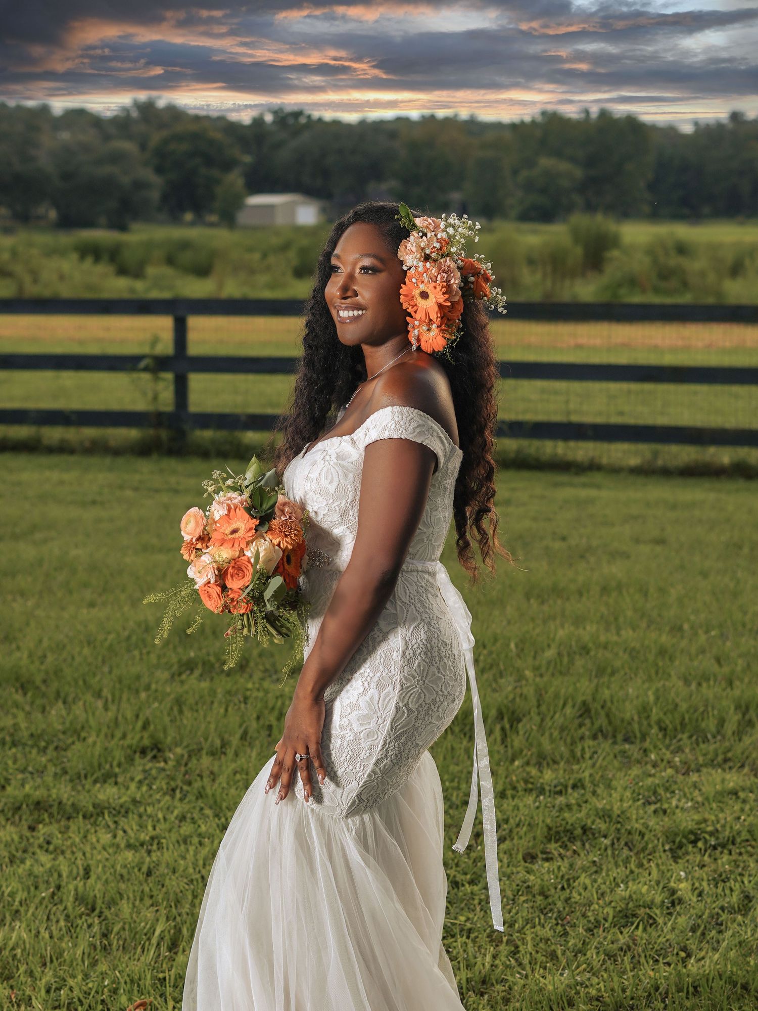 Elegant bridal portrait at sunset with coral flowers and a form-fitting lace wedding gown against a rustic fence backdrop.