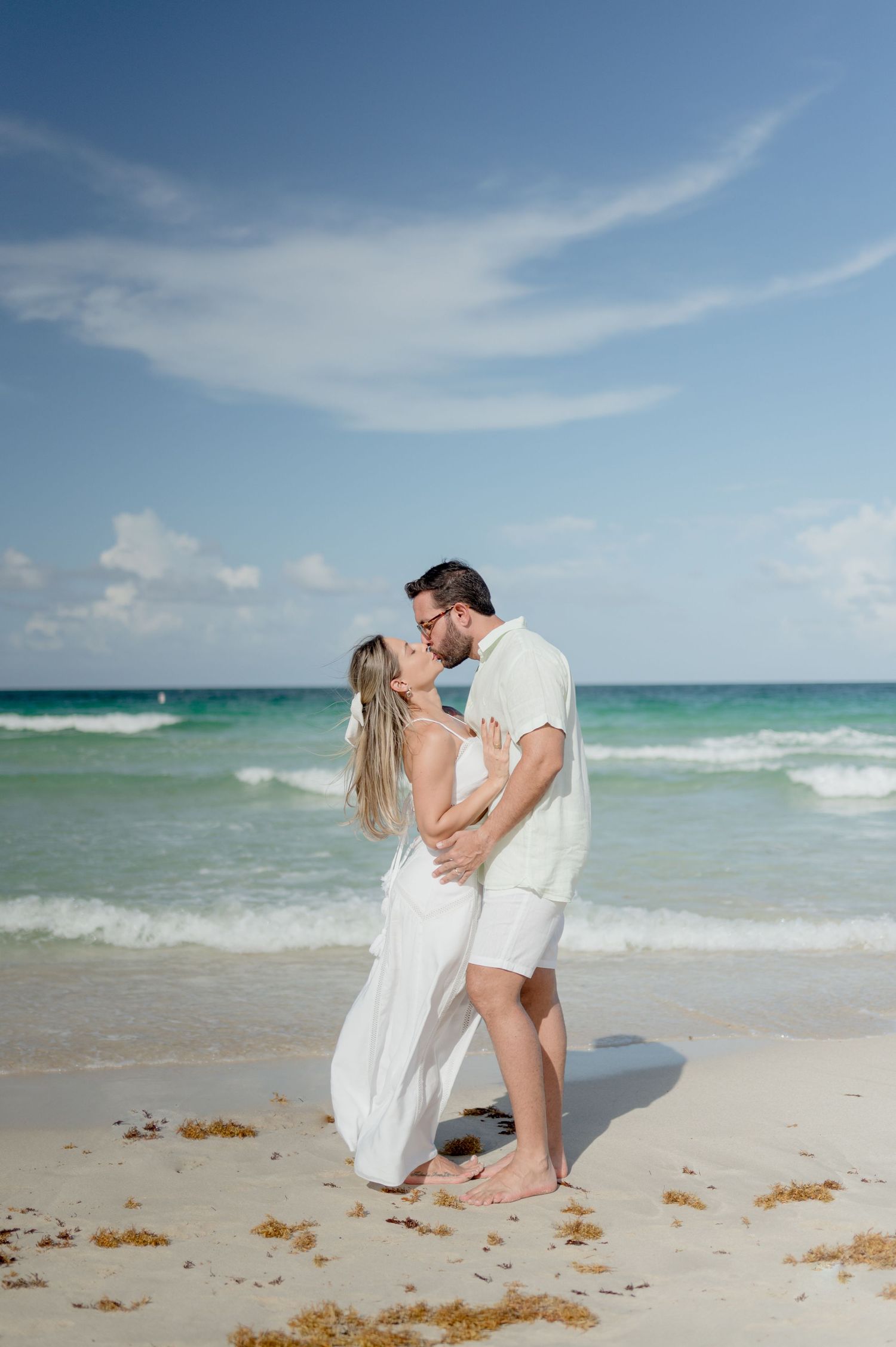 A romantic beach scene with a couple in white clothing embracing on the sand with turquoise waters and blue sky in the background.