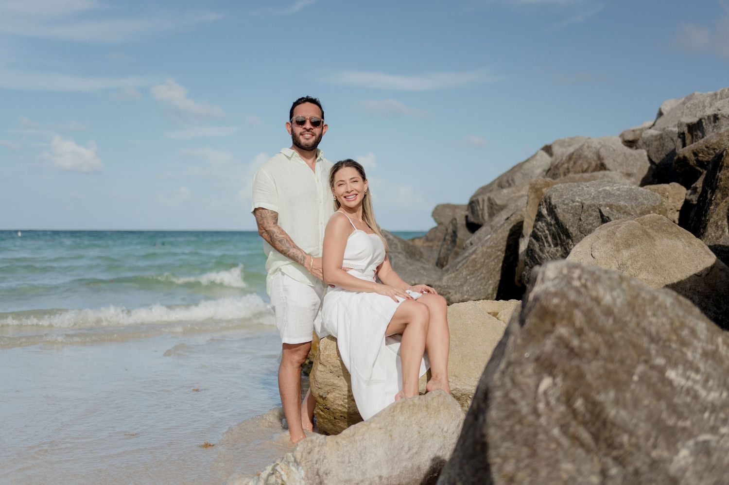 A couple sits together on rocky coastal outcropping overlooking a turquoise ocean on a sunny day.
