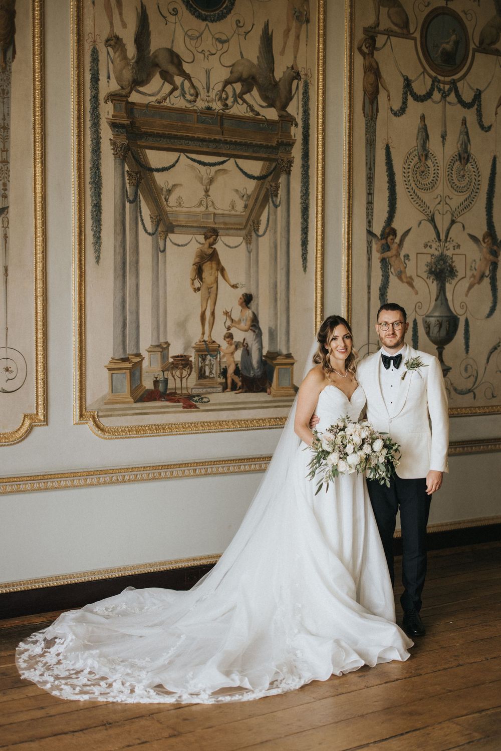 A couple poses in formal wedding attire against an ornate wall with classical artwork and architectural details.