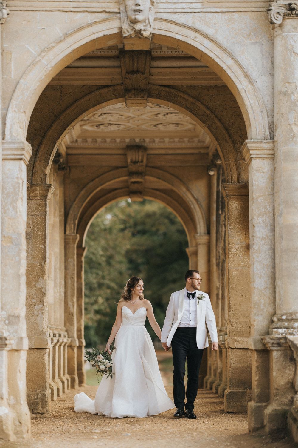A romantic couple in wedding attire poses under a stone archway colonnade with garden views.