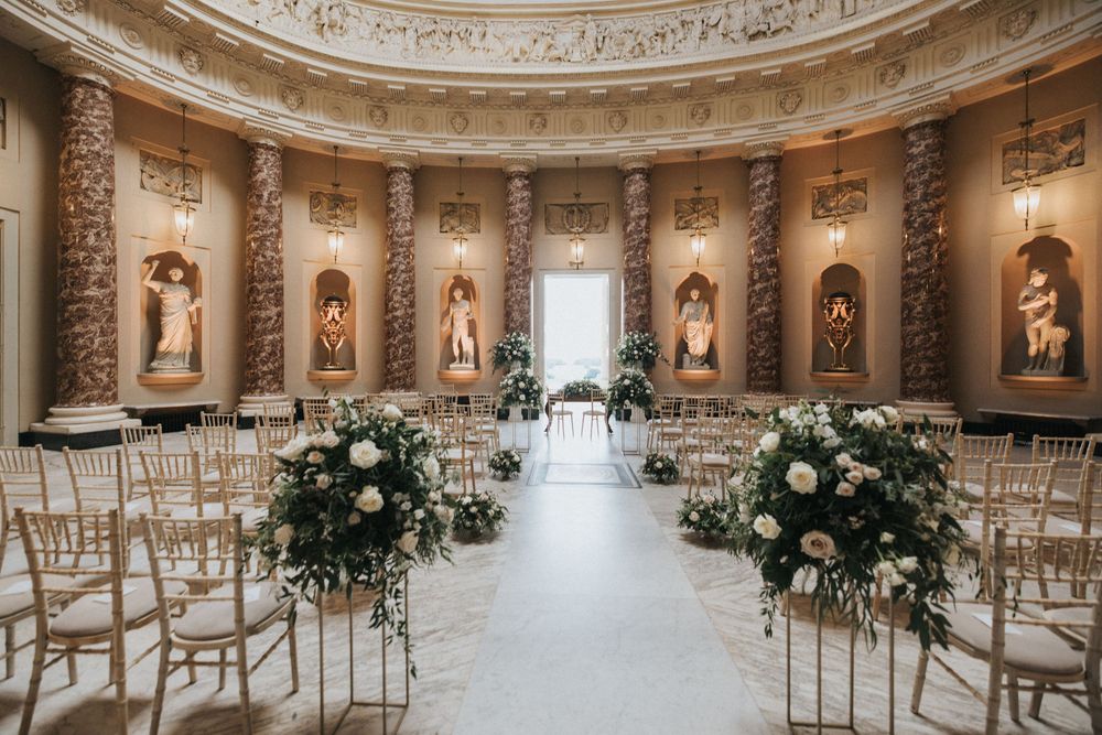 Elegant wedding ceremony setup in circular marble room with statues and floral arrangements along golden chairs.