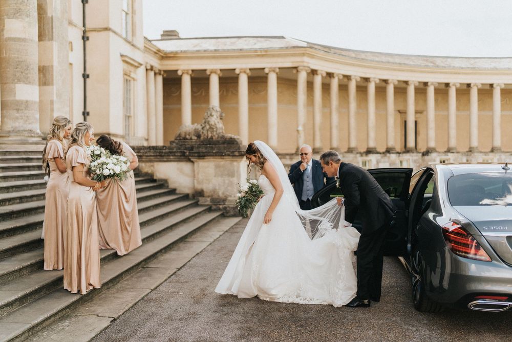 A wedding couple poses for photos along a grand colonnade with classical architecture and pillars in warm sunlight.
