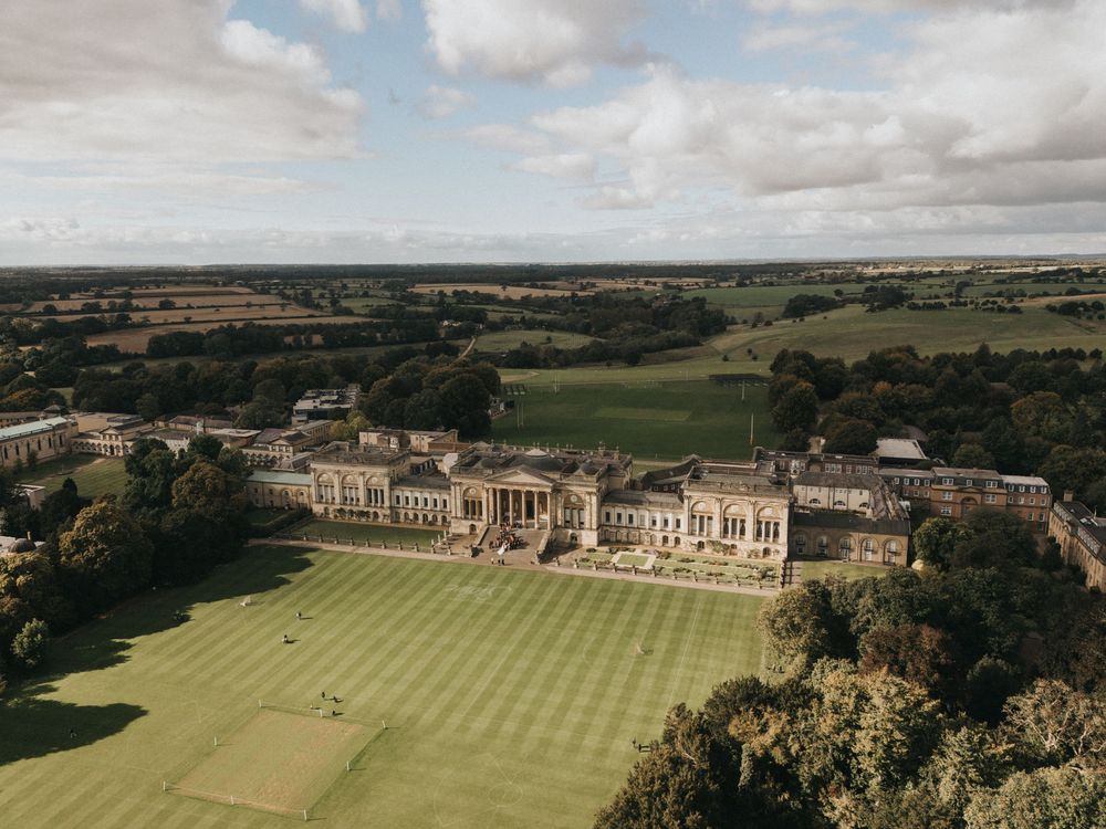 Aerial view of a grand historic building with manicured cricket grounds and rolling countryside in the background.