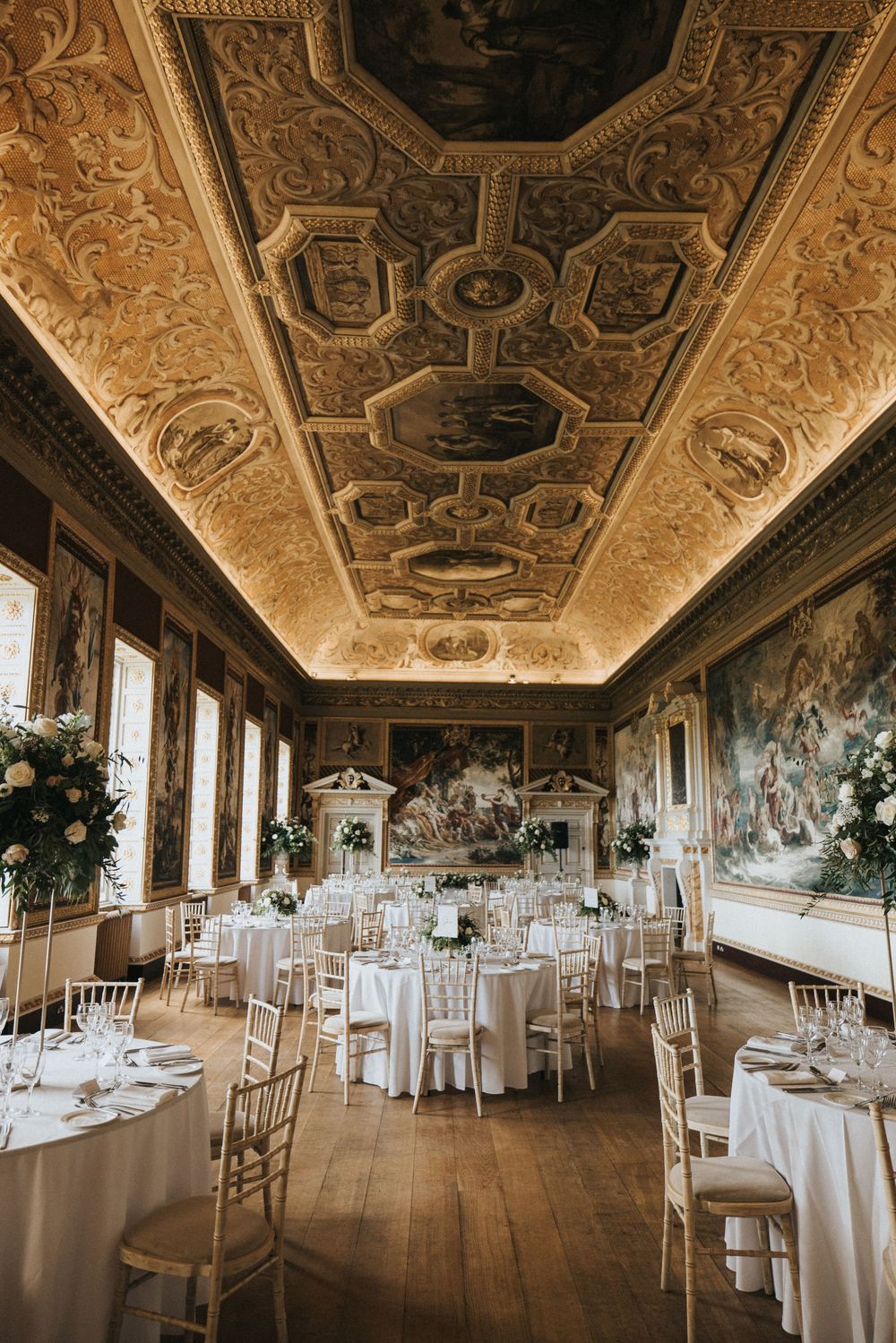 Elegant wedding reception venue with ornate painted ceiling, white round tables, and gold chiavari chairs in grand hall.