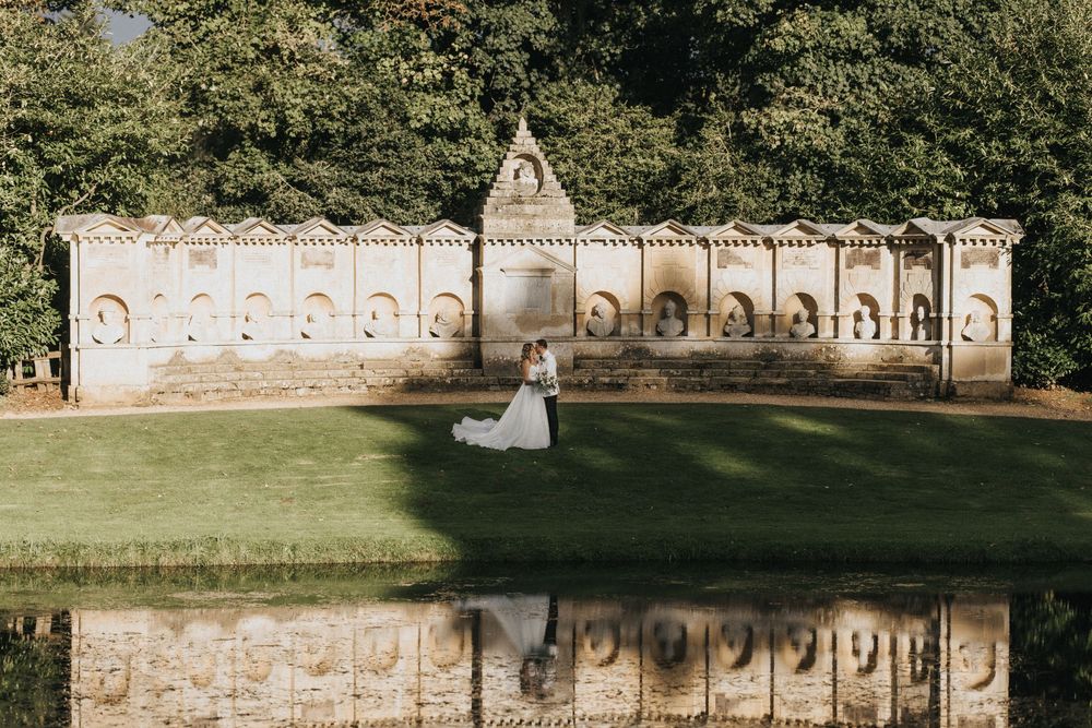 A wedding scene with a stone castle wall in the background reflecting in a pond on a sunny day in a garden setting.