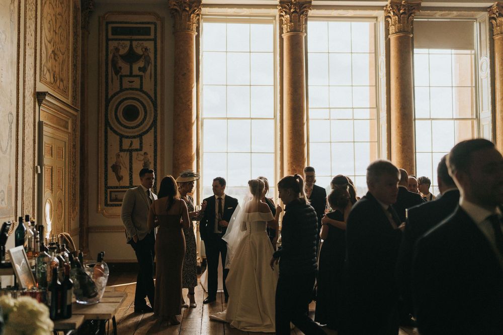 Silhouettes of wedding guests gathered in an elegant room with tall windows and ornate architectural details in warm light.