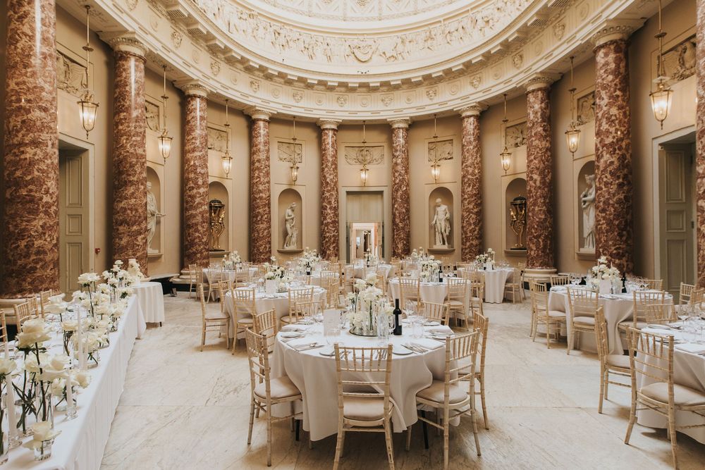 Luxurious circular ballroom with ornate columns and classical statues arranged for a formal reception with round tables and chairs.