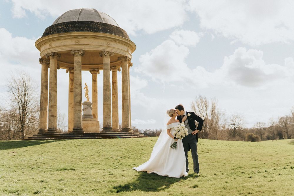 Wedding couple stands near a classical domed rotunda temple structure on a grassy hilltop under cloudy skies.