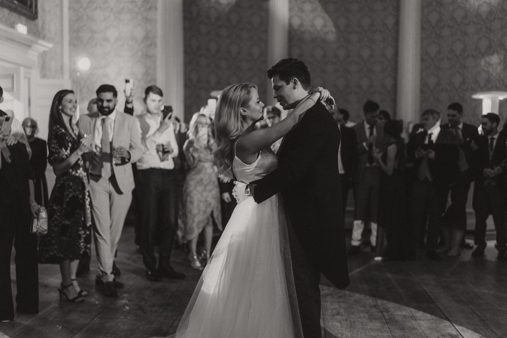 Black and white photo of elegant first dance at wedding reception surrounded by guests watching.