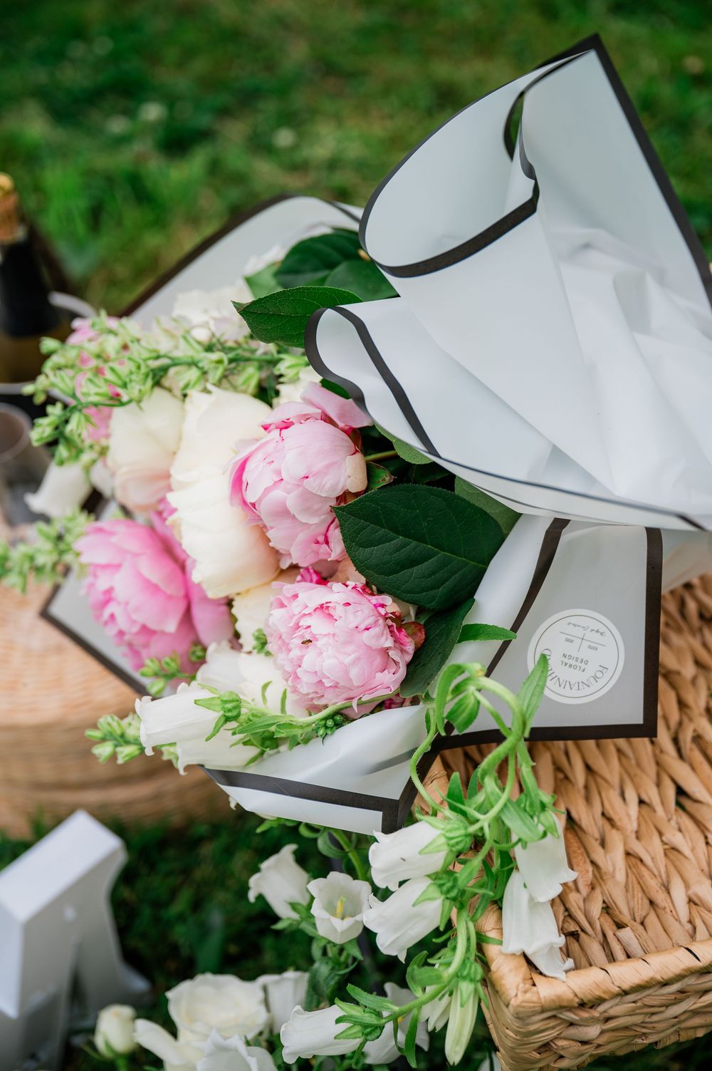 A beautiful bouquet of pink and white peonies wrapped in elegant grey paper sits in a woven basket on grass.