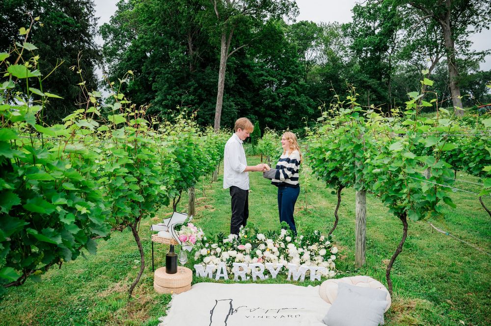 Two people stand together in a lush garden path surrounded by greenery and white floral arrangements.