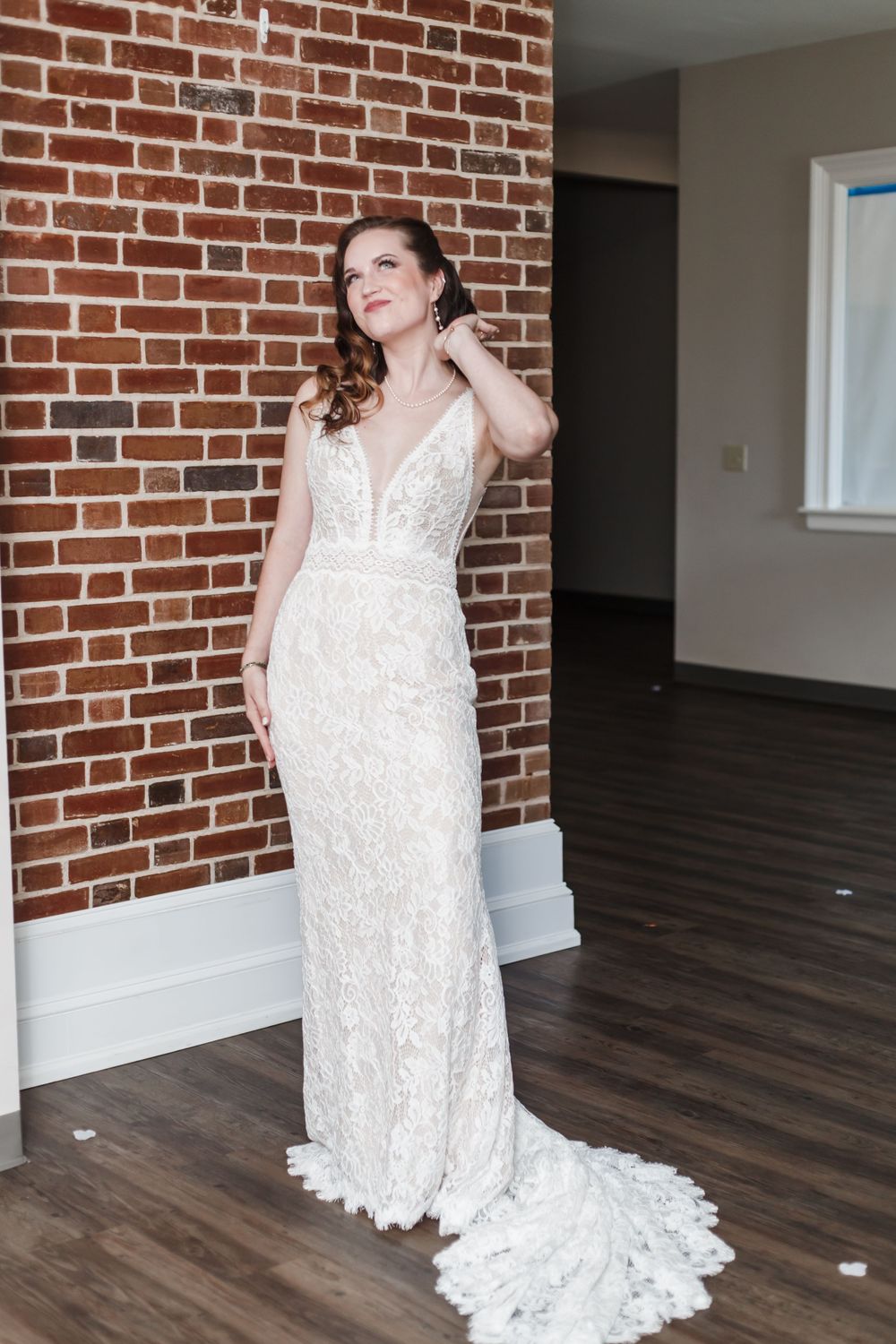 A bride in an elegant white lace wedding gown with a plunging neckline poses against a brick wall backdrop.