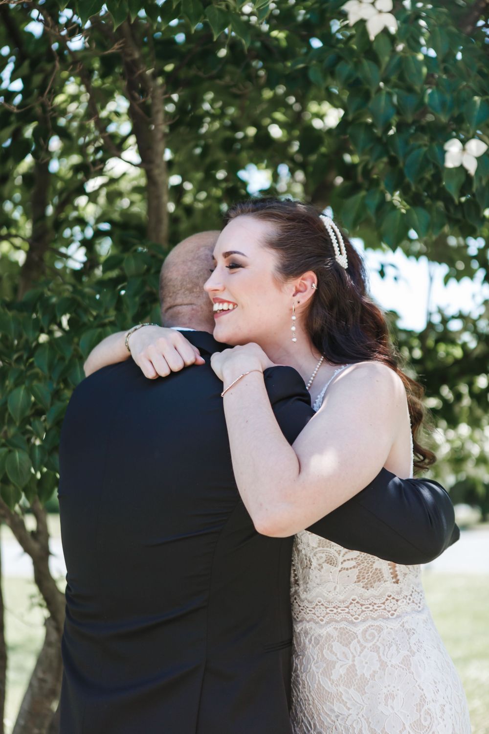 A touching wedding moment as the bride in a lace dress shares an emotional embrace outdoors under green foliage.