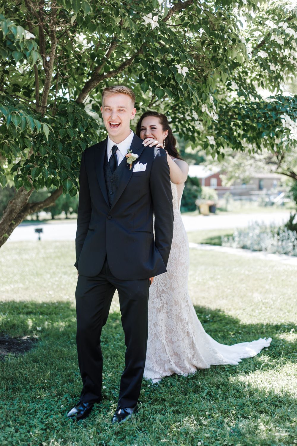 A playful wedding moment captured as a bride peeks out from behind her groom under a leafy tree.