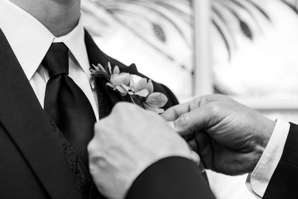 Black and white close-up of someone pinning a boutonniere flower on a suit lapel.