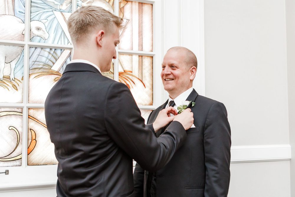 Two men in formal attire adjust each other's boutonnieres before a wedding ceremony.