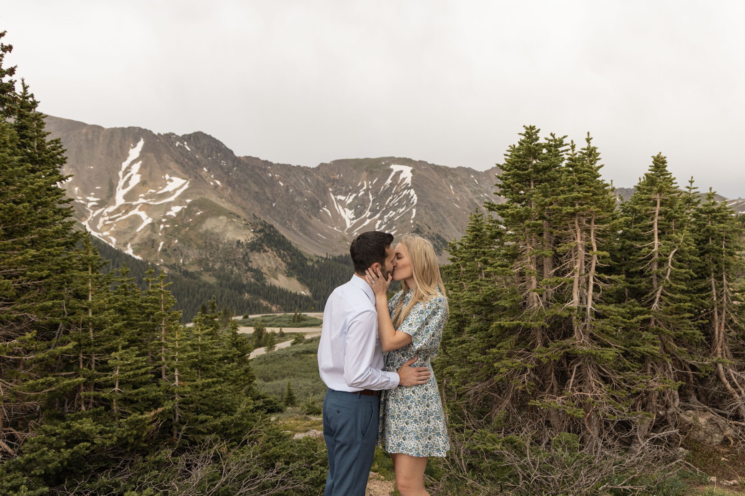 A couple embraces while standing on a mountain overlook with snow-capped peaks and evergreen trees in the background at Loveland Pass