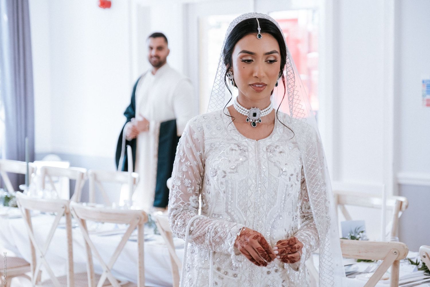 A bride wearing a white lace wedding dress sits at a bright modern reception table in a light-filled venue.