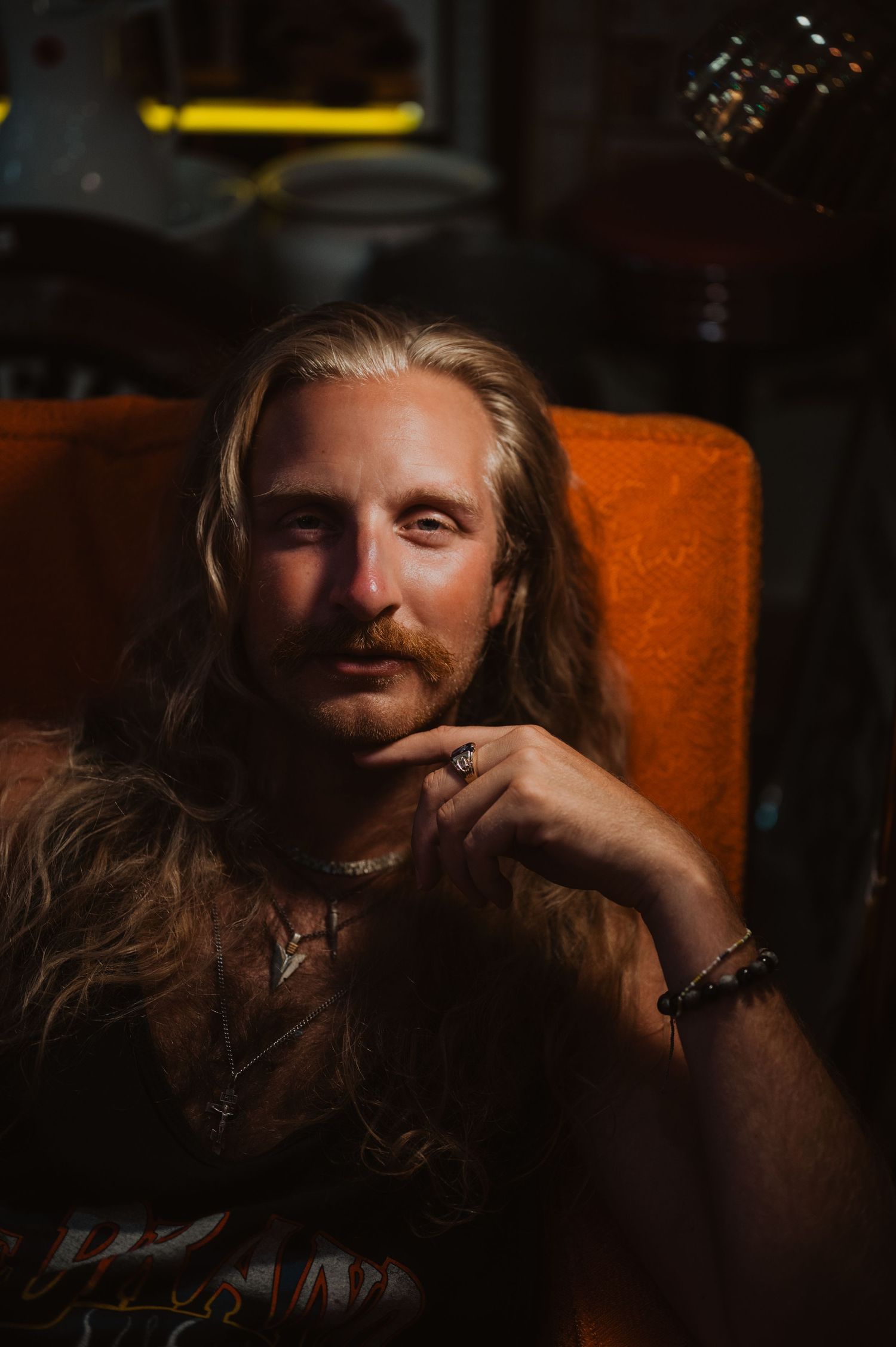Moody portrait of someone with long blonde hair sitting in an orange chair in dim lighting.