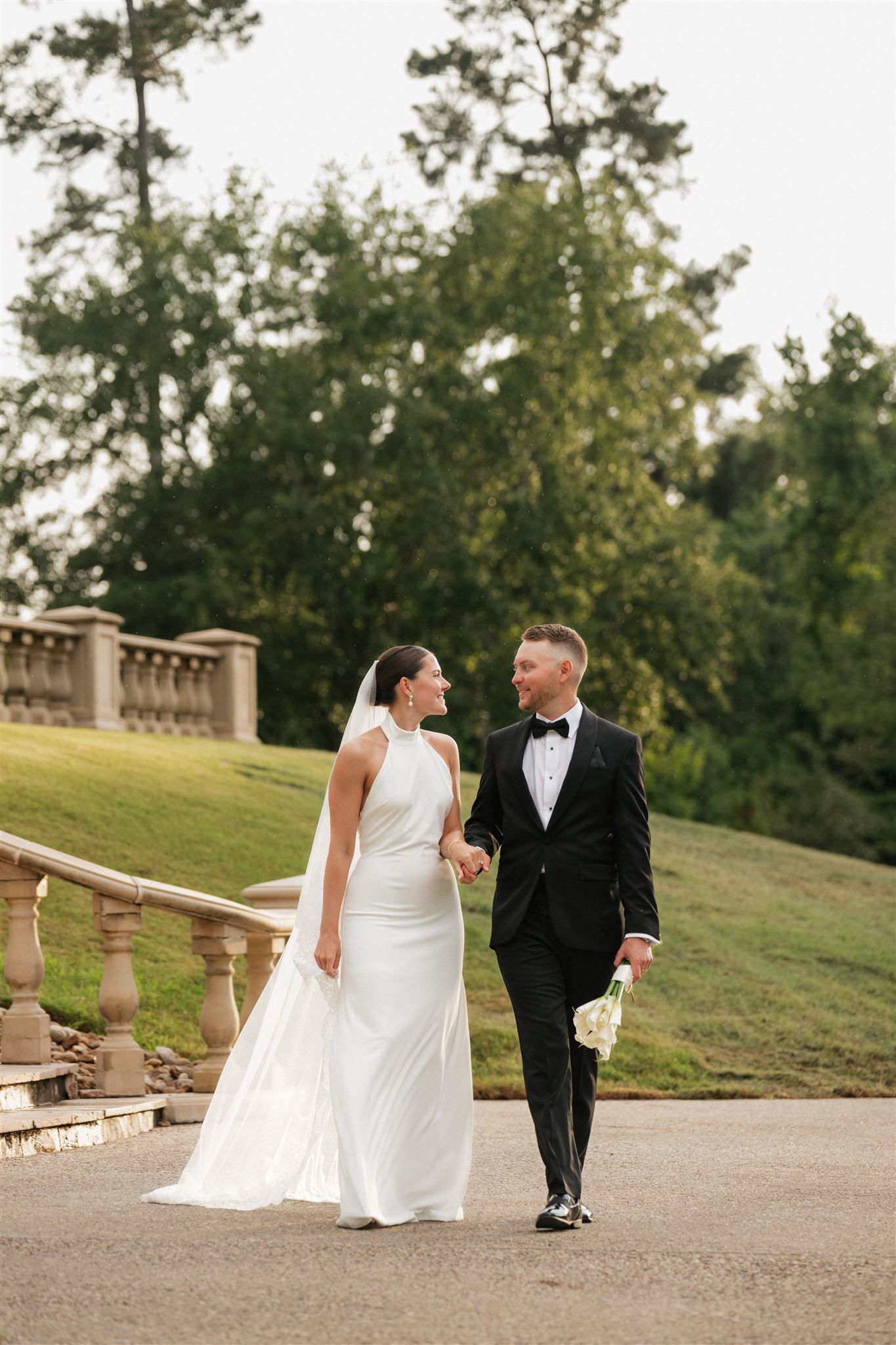 A couple in formal wedding attire walk together along a stone path surrounded by lush greenery and landscaping.