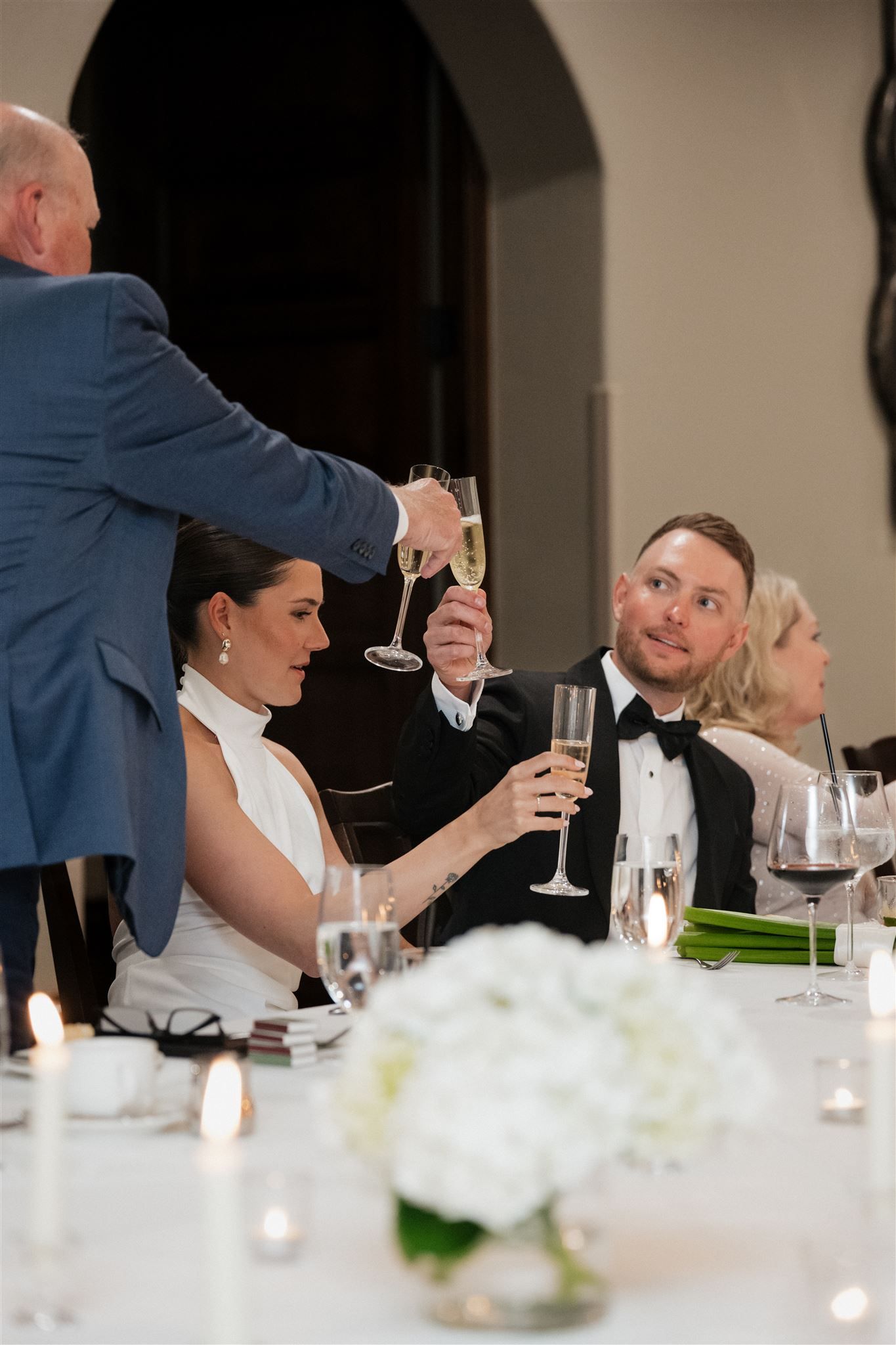 Wedding guests seated at an elegant dinner table share conversation over glasses of wine during a formal reception celebration.