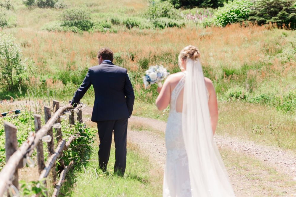 A couple in wedding attire walks hand in hand down a rustic dirt path surrounded by greenery and wooden fencing.