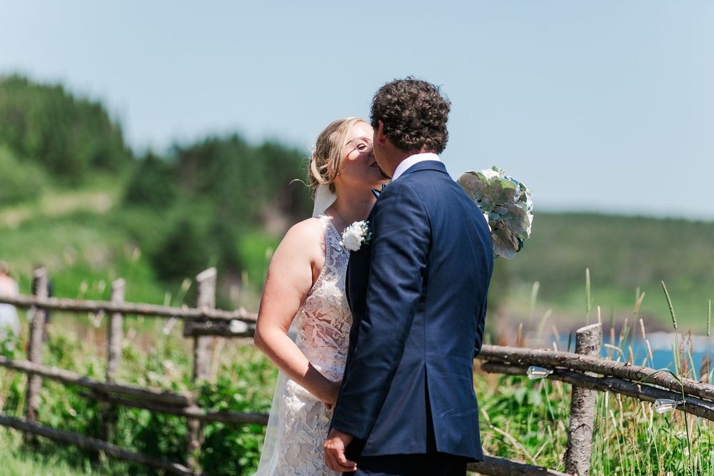A couple shares a romantic kiss on their wedding day in front of a rustic wooden fence with rolling hills in the background.