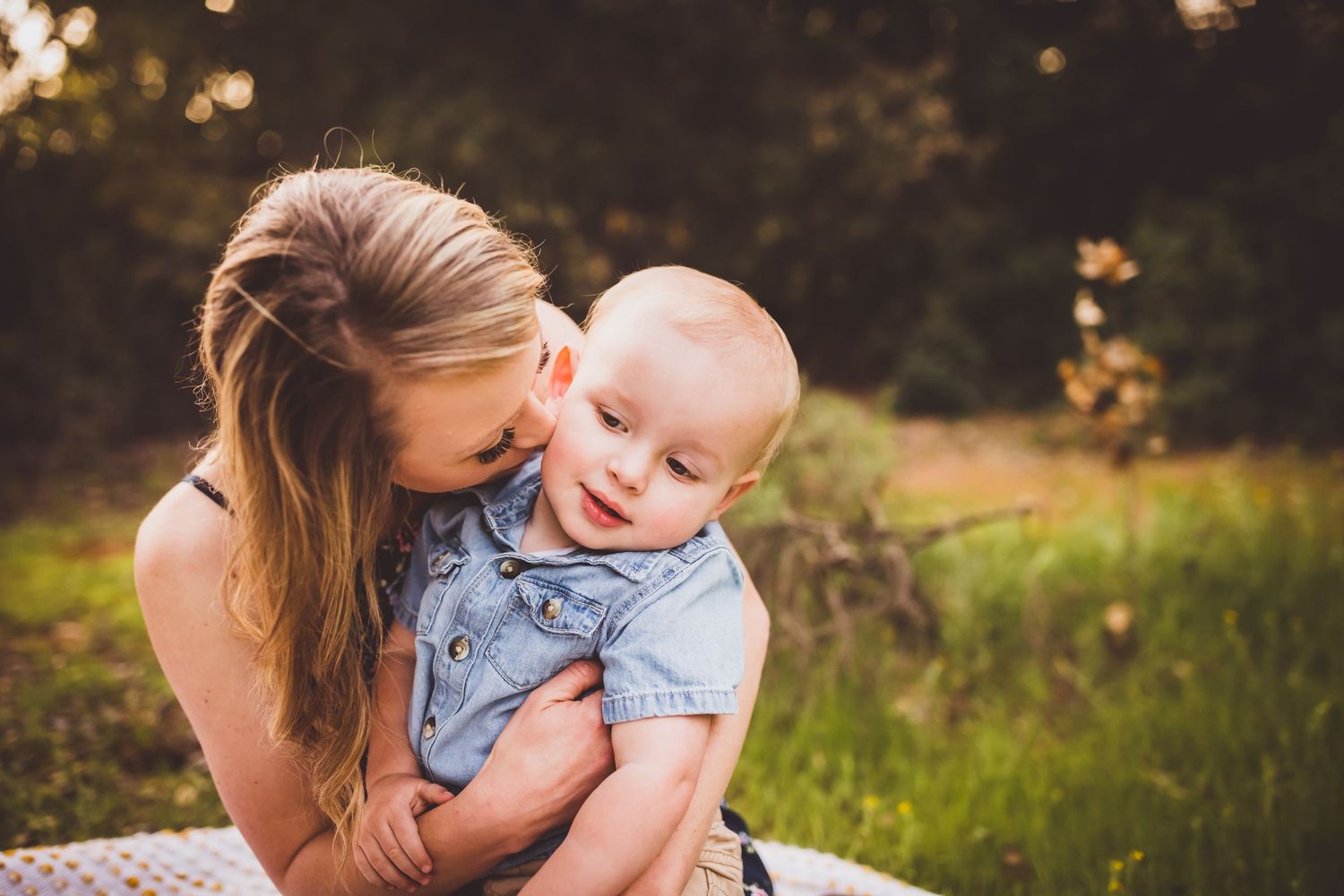 Tender outdoor portrait showing parent and baby sharing joyful moment in sunlit grassy setting.