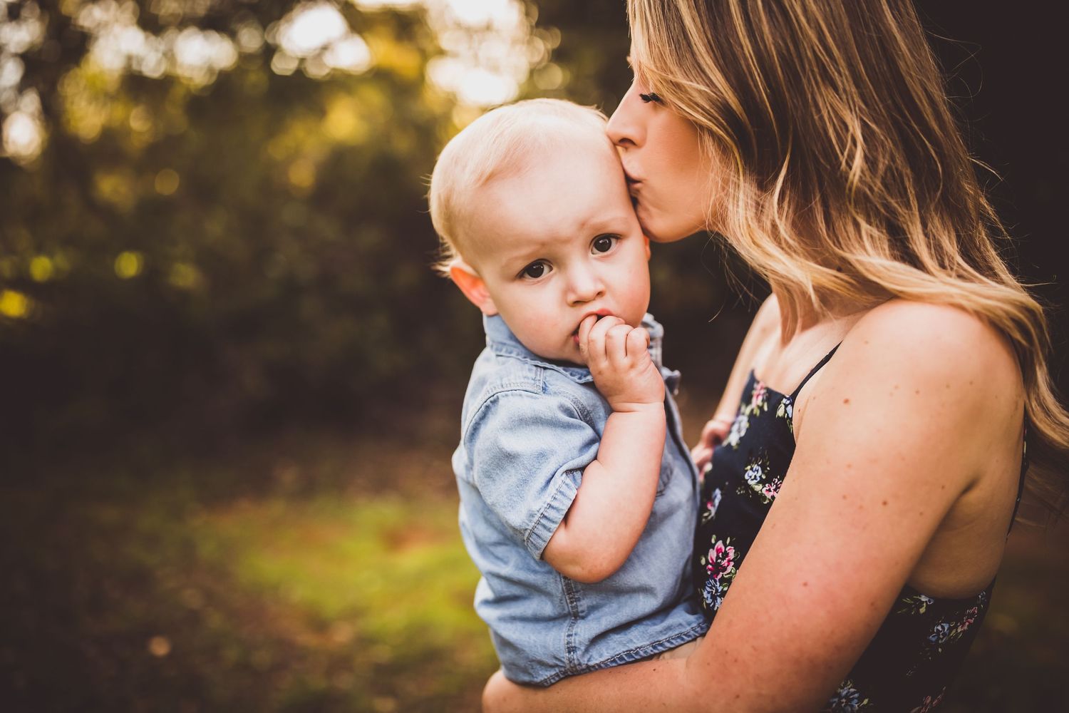 A tender moment captured outdoors between parent and baby in light denim against a soft bokeh sunset background.