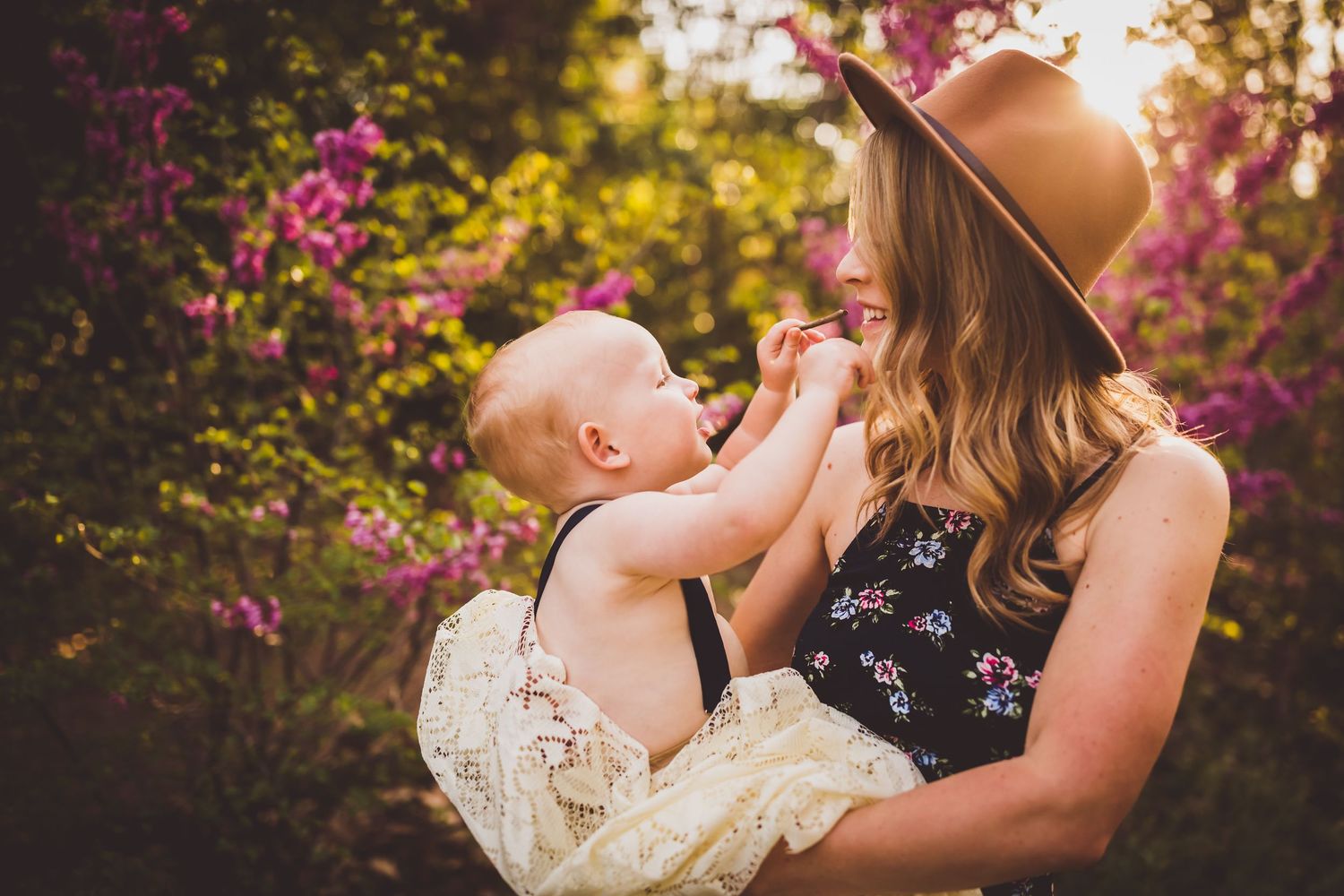 Tender moment captured between mother and baby outdoors with sunlit flowers and greenery in the background.