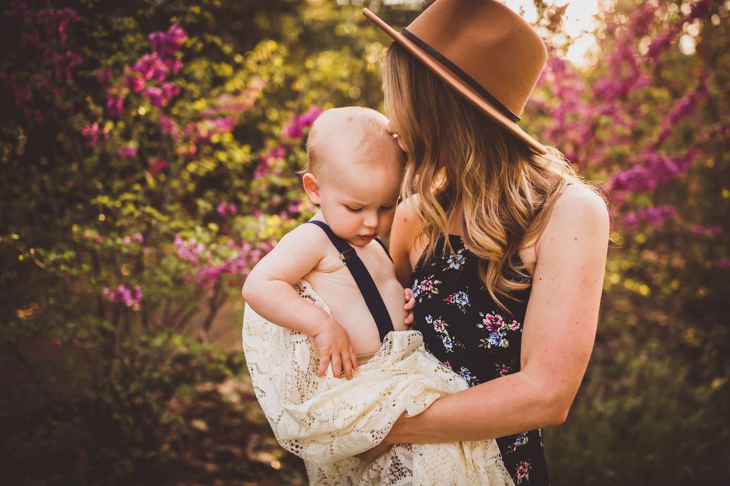 Intimate outdoor portrait with flowering azaleas showing parent wearing hat embracing baby in cream outfit.