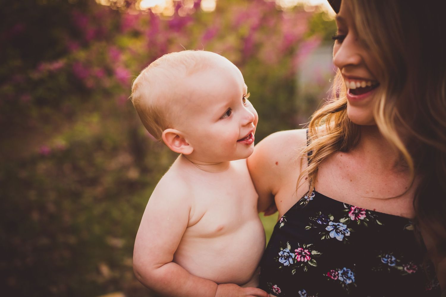 Candid outdoor portrait capturing shared laughter between parent and baby against pink floral background.