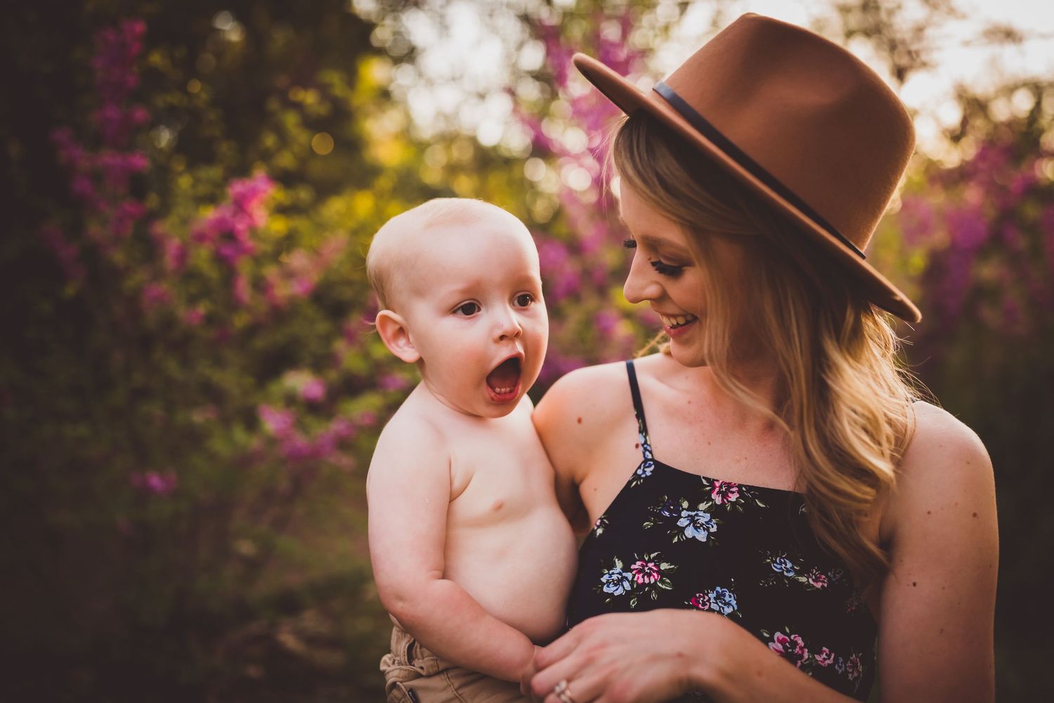 Joyful outdoor portrait with a brown fedora hat against flowering pink bushes.