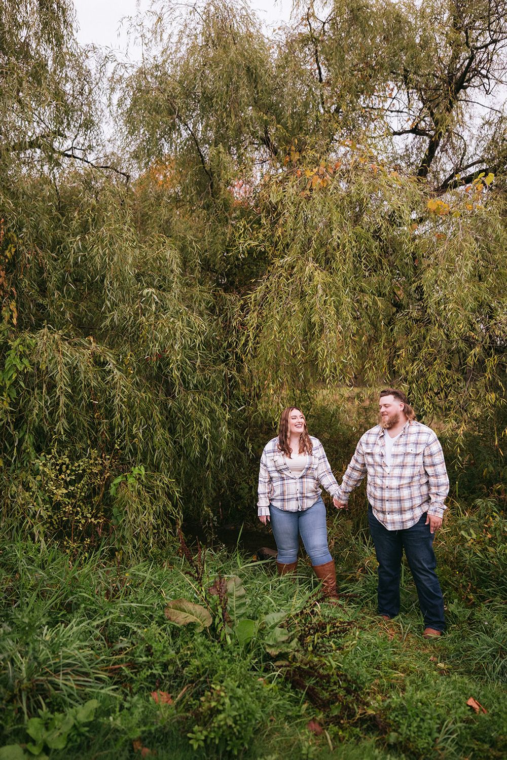 A couple holds hands while standing beneath drooping willow tree branches in autumn.