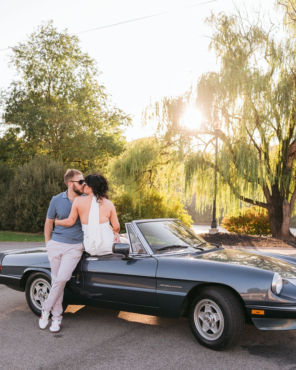 Classic convertible sports car parked under willow trees during golden hour sunset.