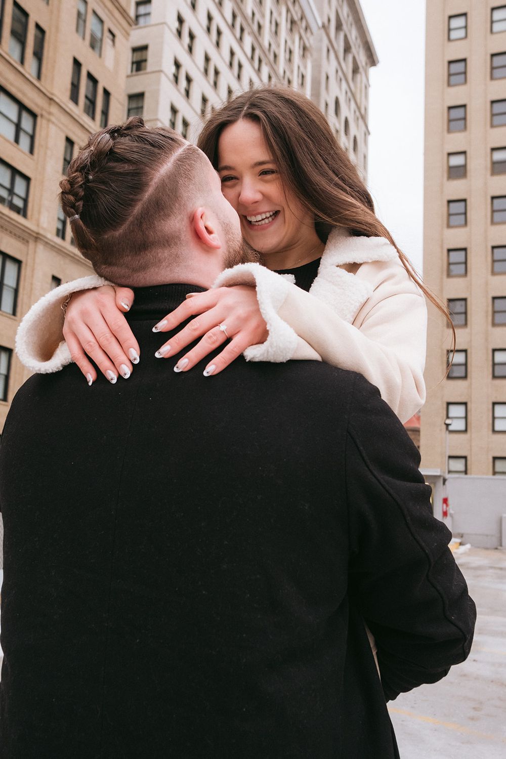 Couple embracing in front of urban buildings with Pittsburgh skyline in background.