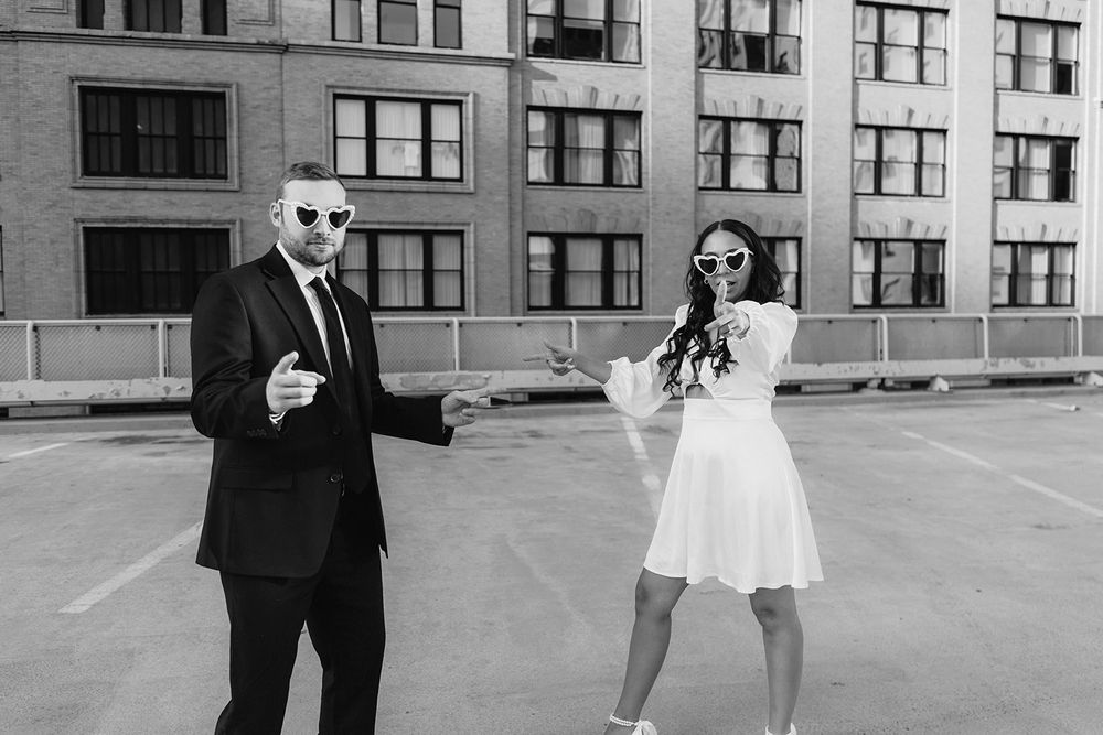 A couple in formal attire pose playfully on an urban rooftop in black and white.