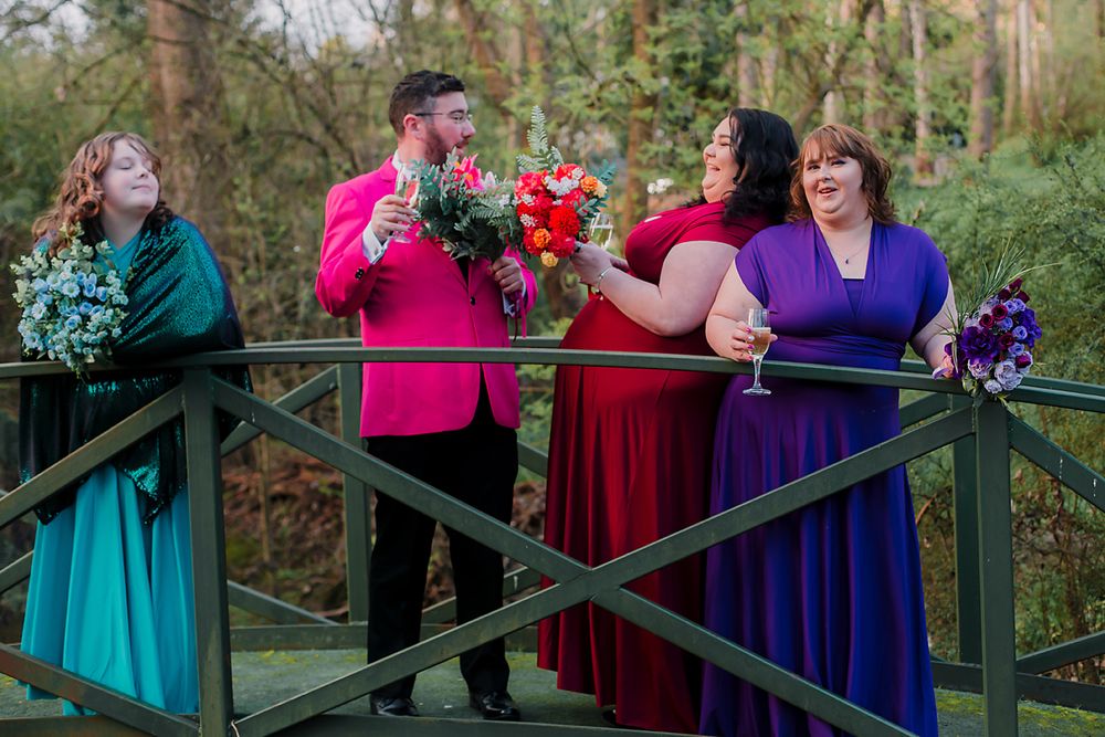 A group of colorfully dressed bridal party members pose together on a wooden bridge in an outdoor setting.