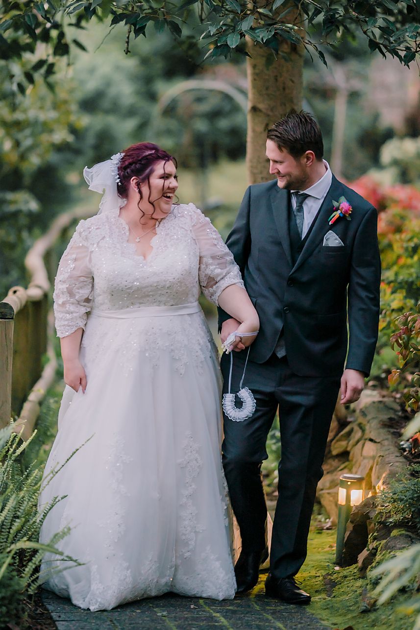 Newlywed couple walking together in garden setting with bride in lace dress and groom in dark suit.