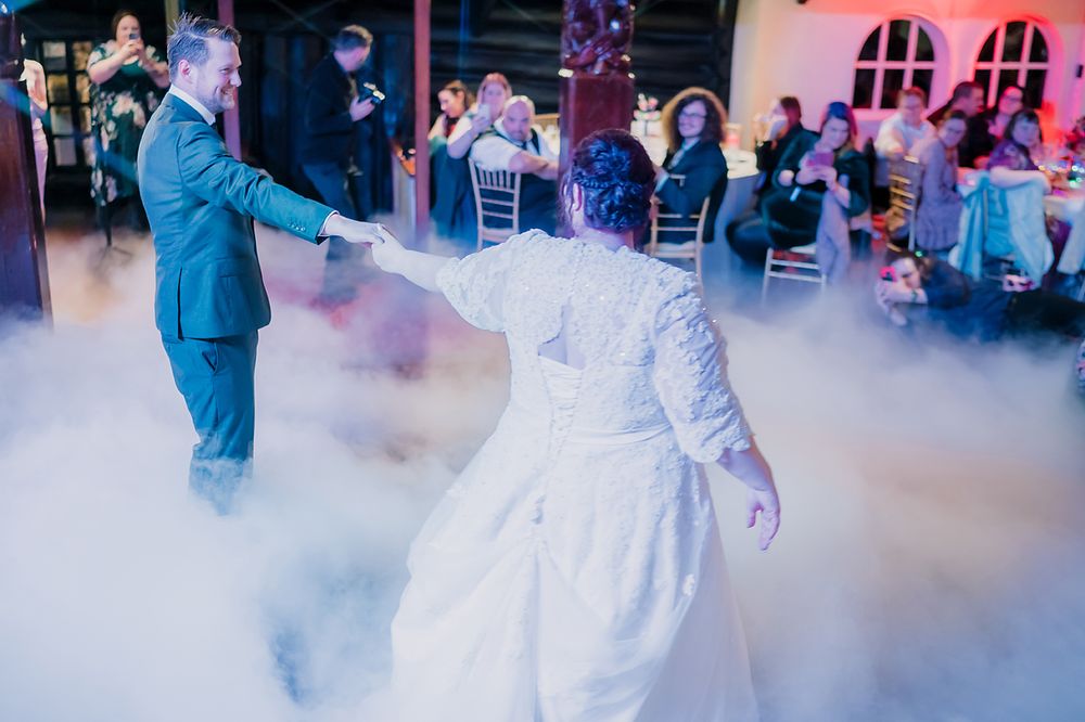 A couple sharing their first dance as newlyweds on a misty dance floor with purple lighting at their wedding reception.