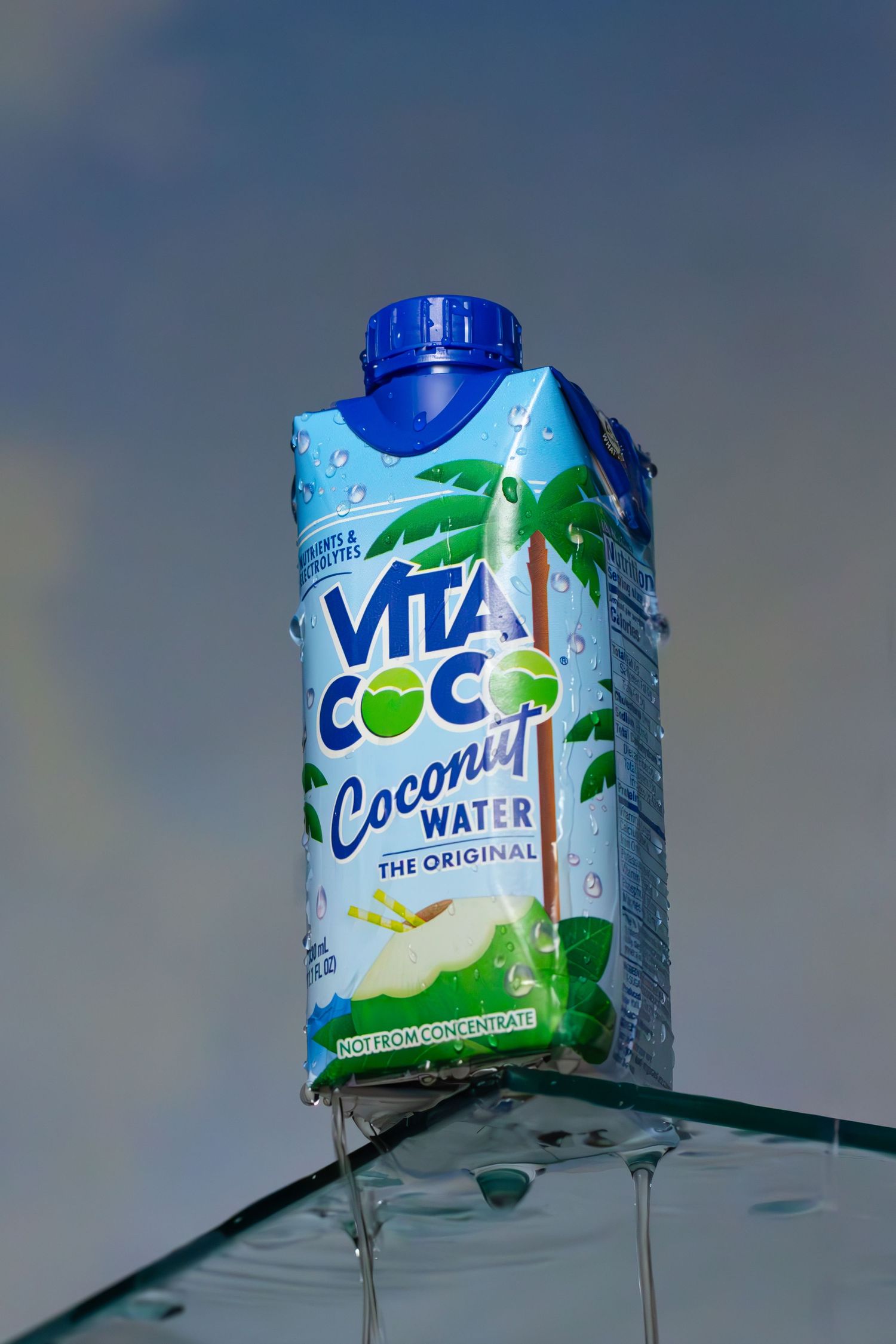 A carton of Vita Coco coconut water with palm tree graphics sitting on clear glass surface against blue sky background.