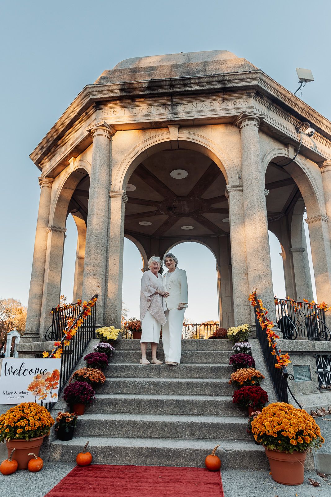 A couple stands in a grand stone gazebo decorated with orange mums and pumpkins for their autumn wedding.