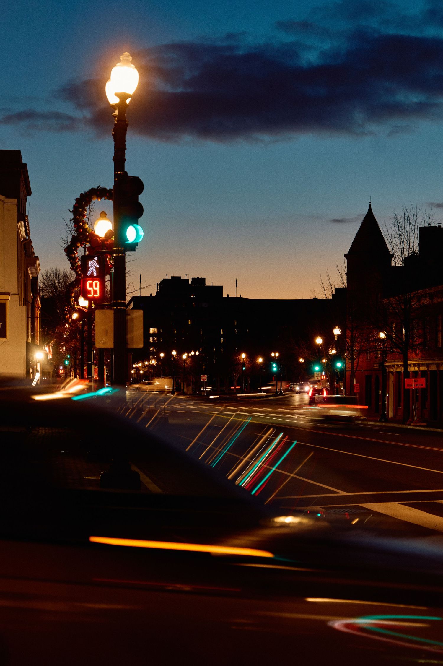 A street at dusk with illuminated lampposts and traffic lights creating streaks of color against the darkening sky.